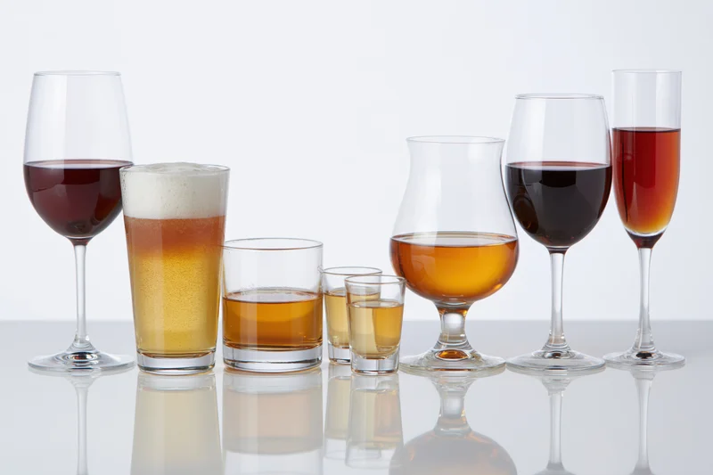 Assorted drink glasses filled with various alcoholic beverages arranged in a row on a reflective white surface