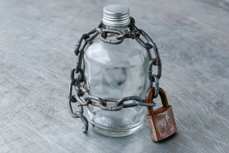 Empty glass liquor bottle wrapped in metal chain and secured with padlock on textured gray surface