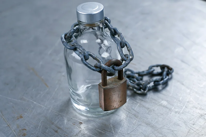Empty glass liquor bottle secured with heavy metal chain and padlock on scratched steel surface