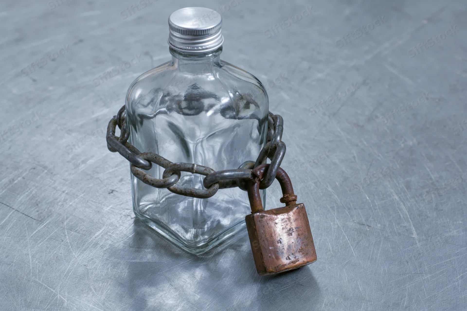 Empty glass liquor bottle secured with a heavy metal chain and padlock on a scratched steel surface