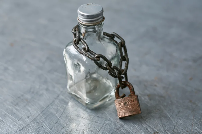 Empty glass liquor bottle secured with heavy metal chain and padlock on textured gray surface