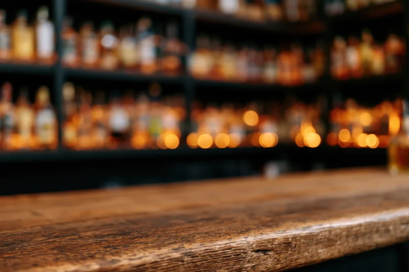 Empty worn wooden bar counter in foreground with blurred liquor bottles on shelves behind