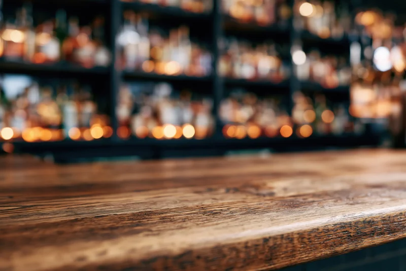 Worn wooden bar counter in foreground with blurred liquor bottles on shelves behind
