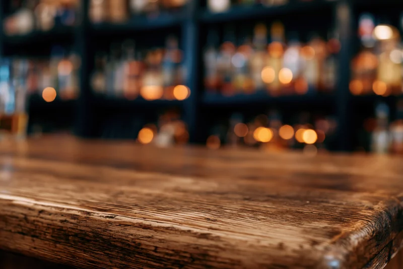 Worn wooden bar counter in focus with blurred liquor bottles and warm lights in background