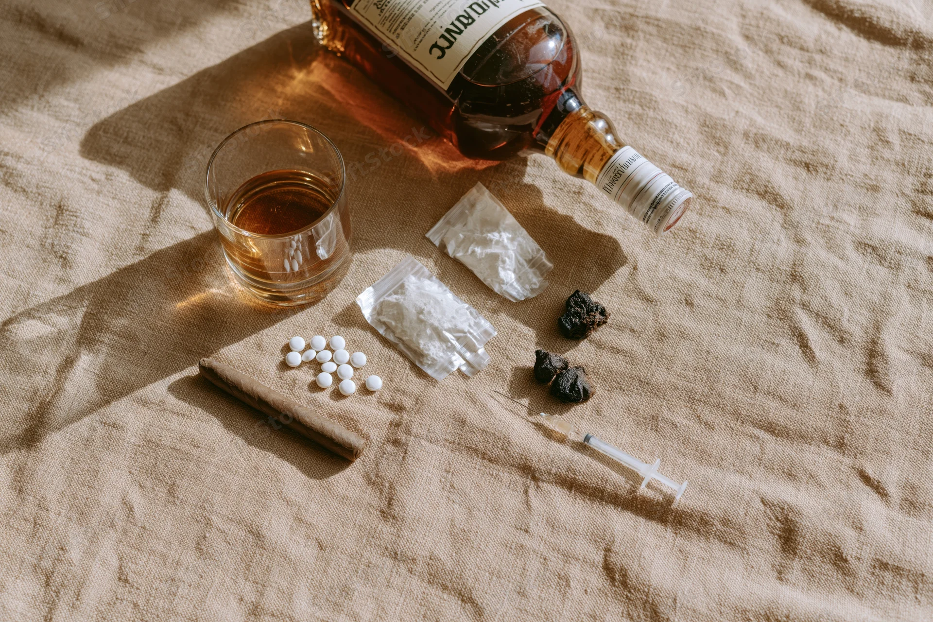 Flat lay of addiction items including whiskey bottle, glass, pills, syringe, and drugs on beige fabric