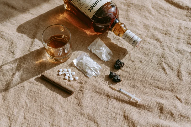 Flat lay of addiction items including whiskey bottle, glass, pills, syringe, and drugs on beige fabric