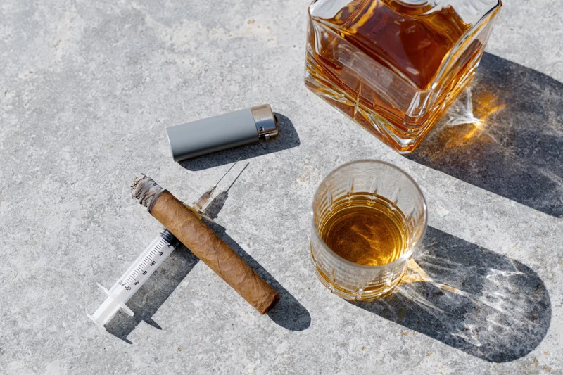 Whiskey glass, cigar, lighter and syringe arranged on concrete surface in flat lay overhead view
