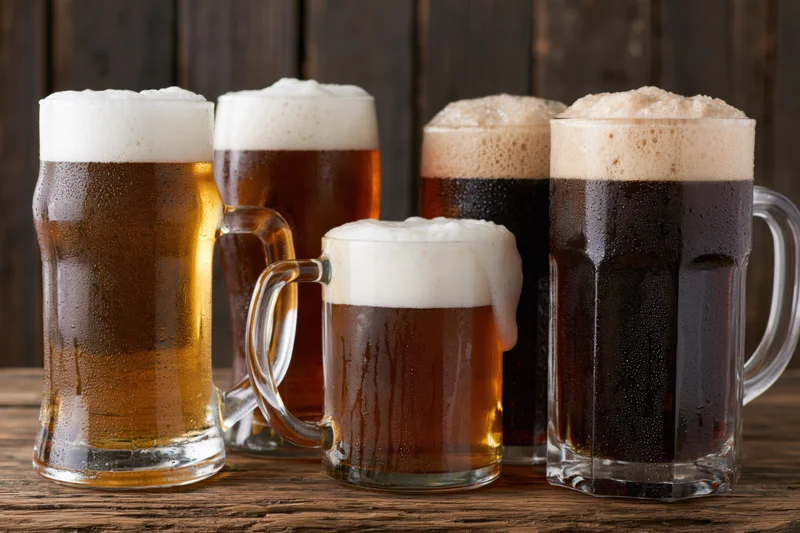 Five beer glasses and mugs with frothy heads arranged on a wooden table