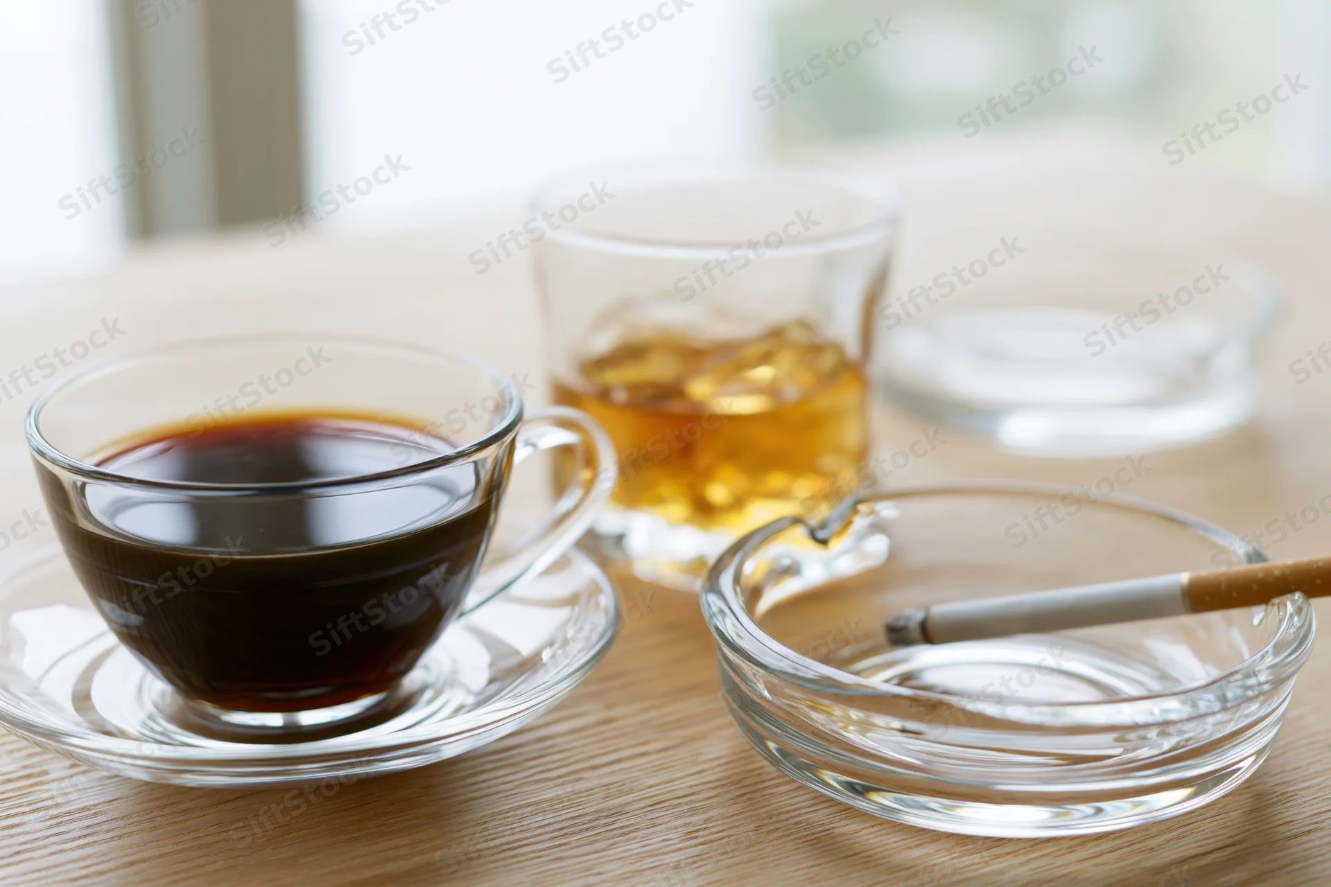 Glass cup of black coffee on saucer beside whiskey glass and ashtray with cigarette on wooden table