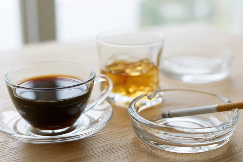 Glass cup of black coffee on saucer beside whiskey glass and ashtray with cigarette on wooden table