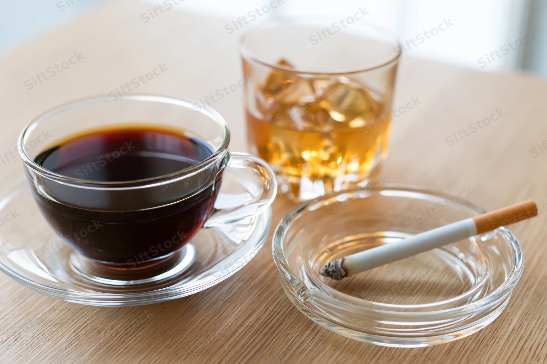 Glass cup of black coffee on saucer beside whiskey glass and cigarette in ashtray on wooden table