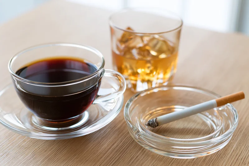 Glass cup of black coffee on saucer beside whiskey glass and cigarette in ashtray on wooden table