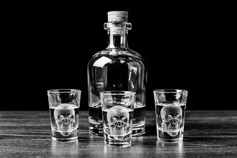 Glass decanter with cork stopper flanked by three skull-etched shot glasses on wooden table
