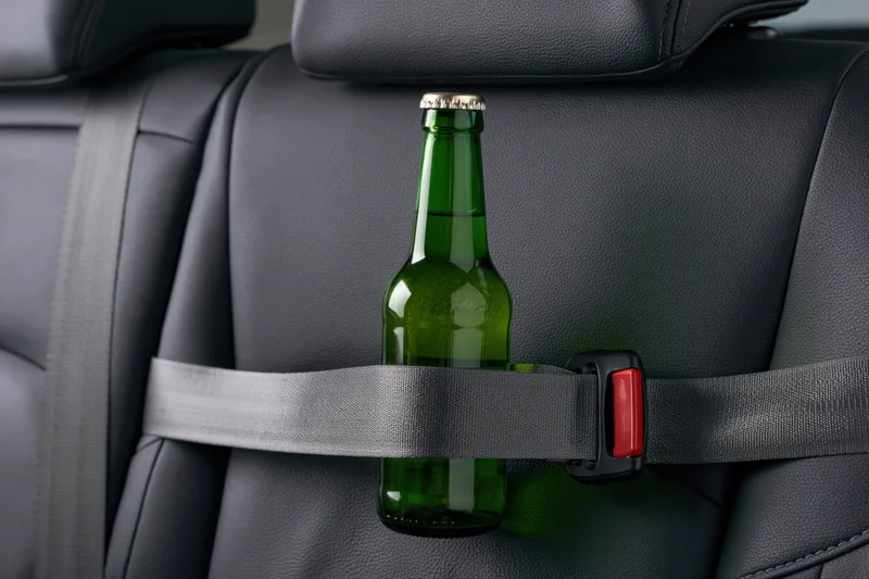 Green glass beer bottle secured with gray seatbelt in car seat interior