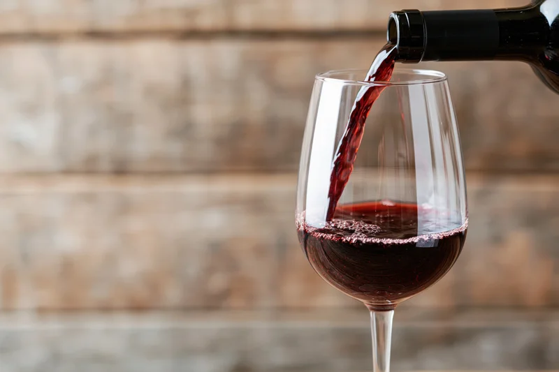 Red wine pouring from a dark bottle into a clear stemmed glass against a wooden background