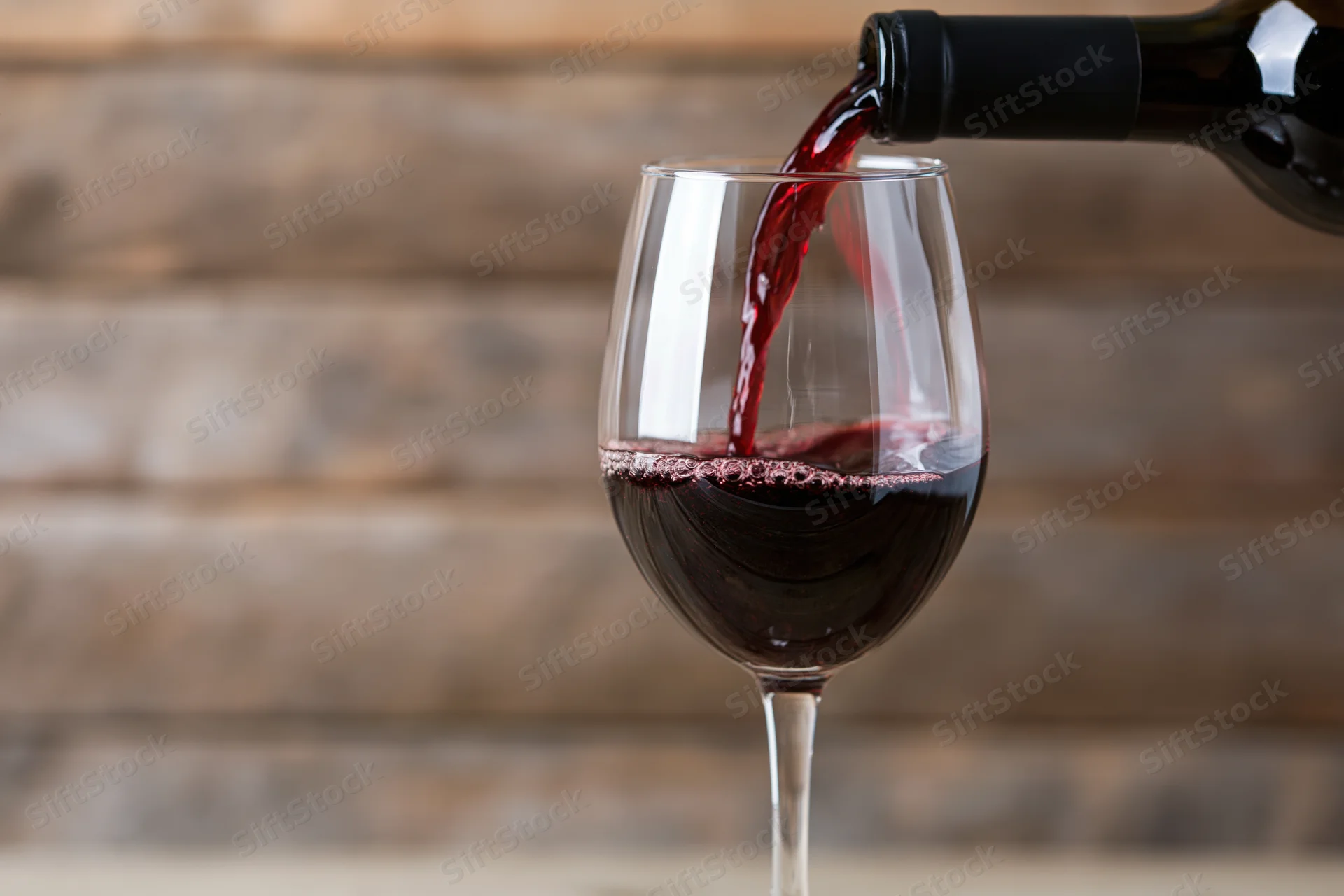Red wine being poured from a dark bottle into a clear stemmed glass against a wooden background
