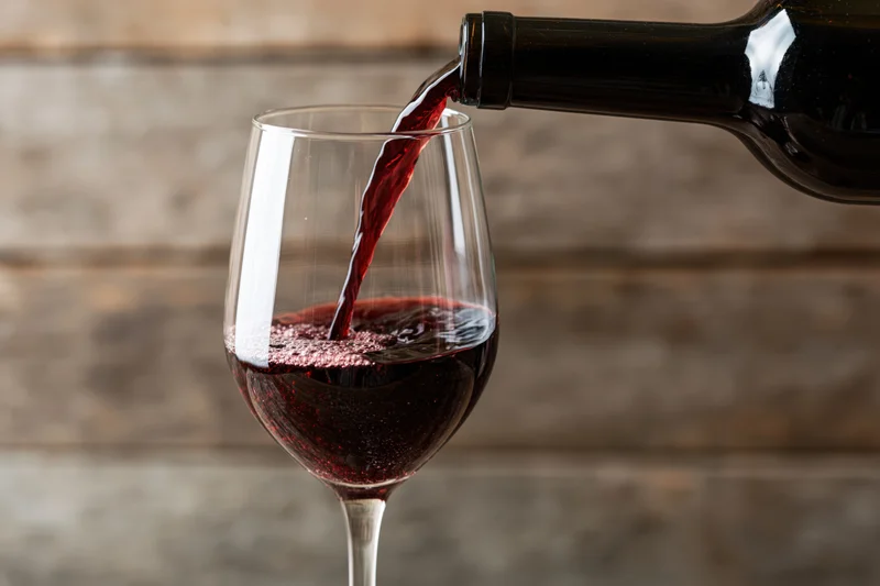 Red wine pouring from a dark bottle into a clear stemmed glass against a wooden background