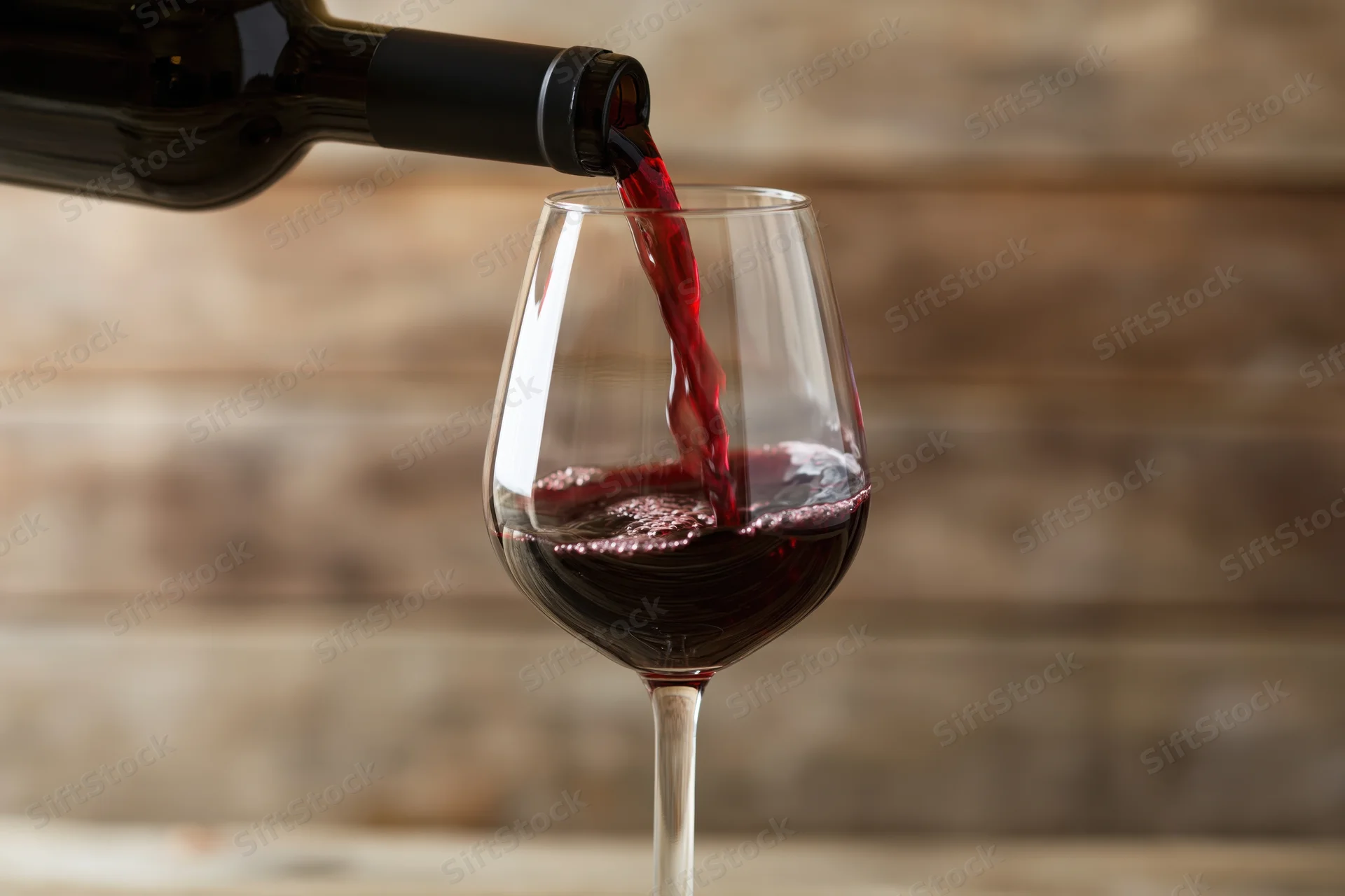 Red wine pouring from a dark bottle into a clear stemmed glass against a wooden background
