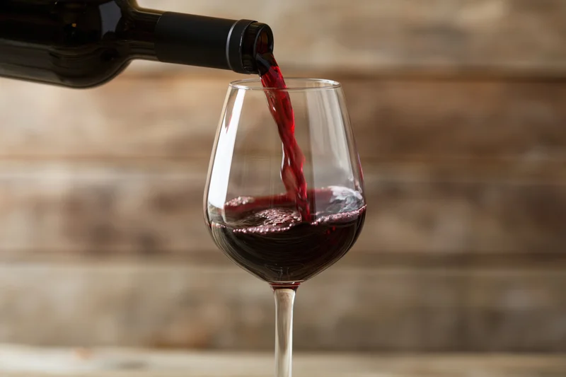 Red wine pouring from a dark bottle into a clear stemmed glass against a wooden background