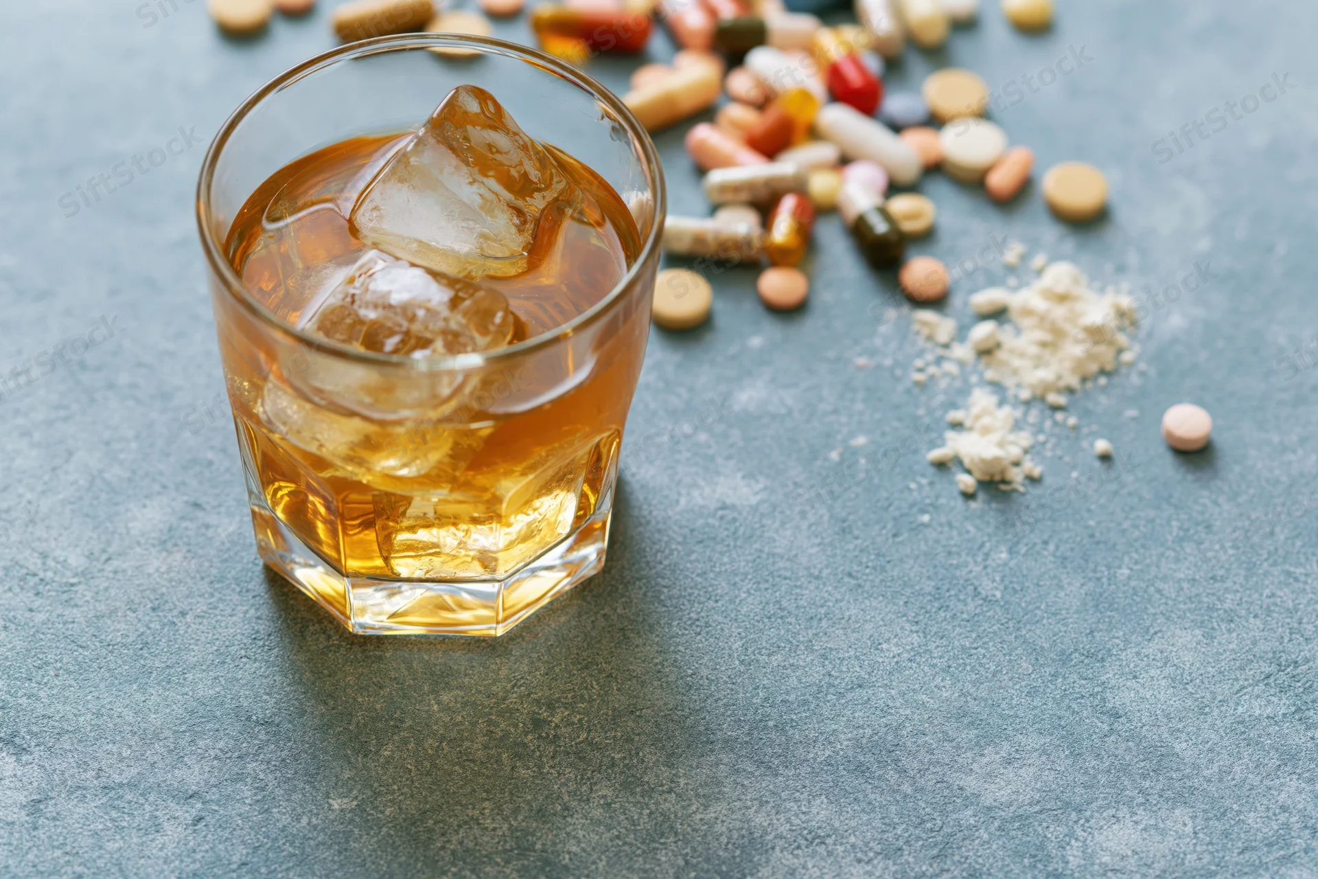 Whiskey in a rocks glass with ice beside scattered pills and powder on a gray surface