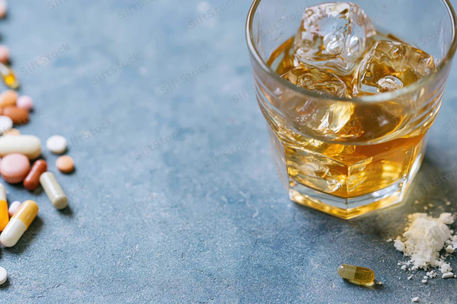 Whiskey in a rocks glass with ice cubes on a blue surface beside scattered pills and powder