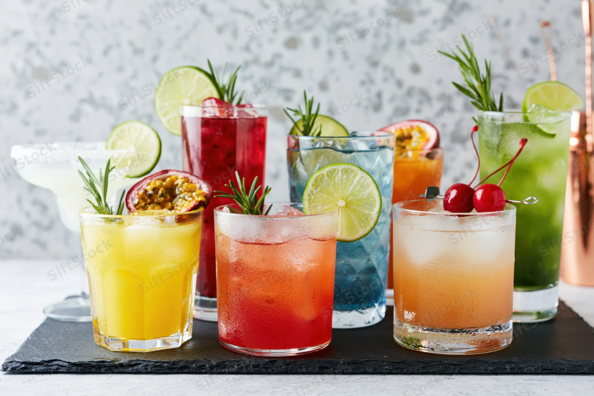 Eight assorted cocktails with fruit and herb garnishes arranged on a dark slate board