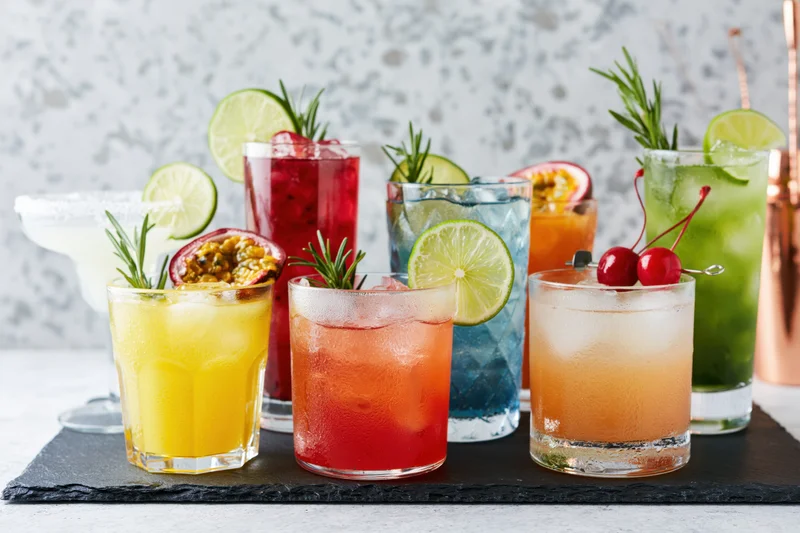 Eight assorted cocktails with fruit and herb garnishes arranged on a dark slate board