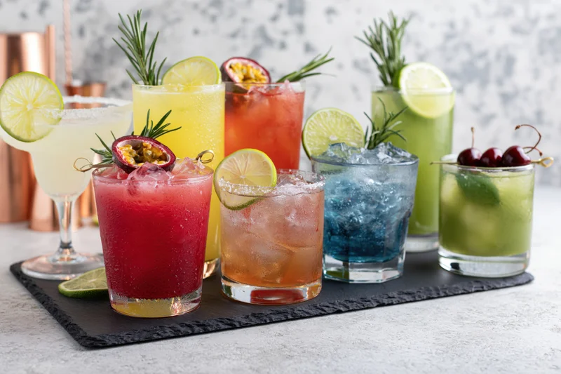 Seven colorful cocktails garnished with fruit and herbs arranged on a dark slate tray on a countertop