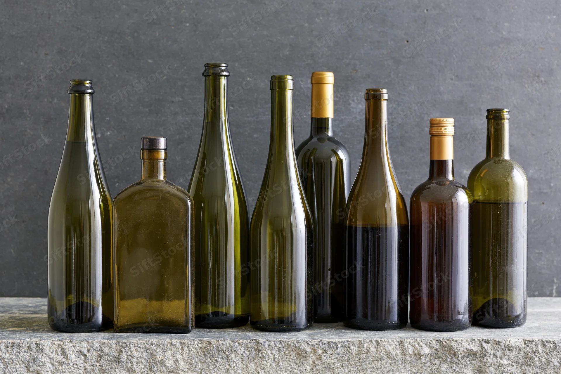 Seven assorted unlabeled liquor and wine bottles arranged in a row on a stone surface against a dark gray background