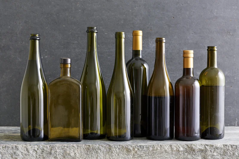 Seven assorted unlabeled liquor and wine bottles arranged in a row on a stone surface against a dark gray background