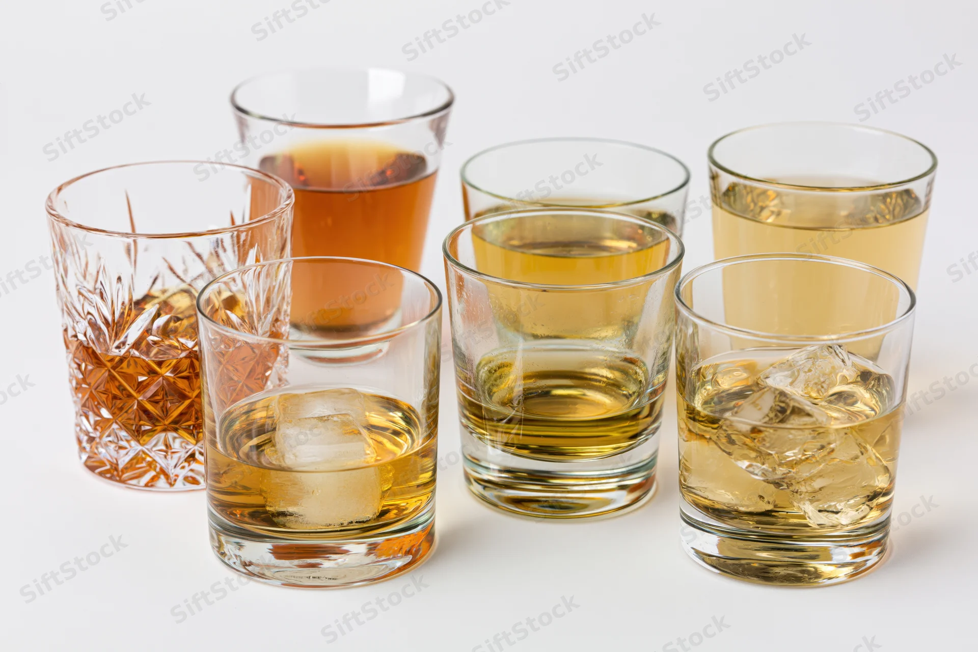 Seven whiskey glasses of varying styles and fills arranged on a white surface with ice and amber liquid