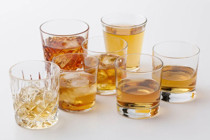 Seven whiskey glasses of varying styles and fills arranged on a white surface