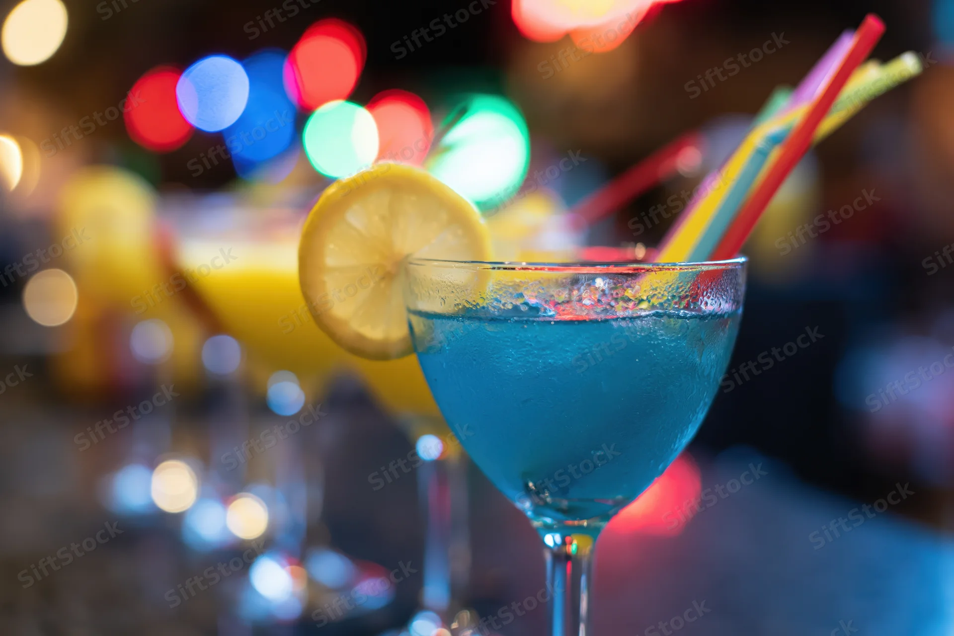 Blue cocktail in a glass with lemon slice and rainbow straw on a bar counter at night