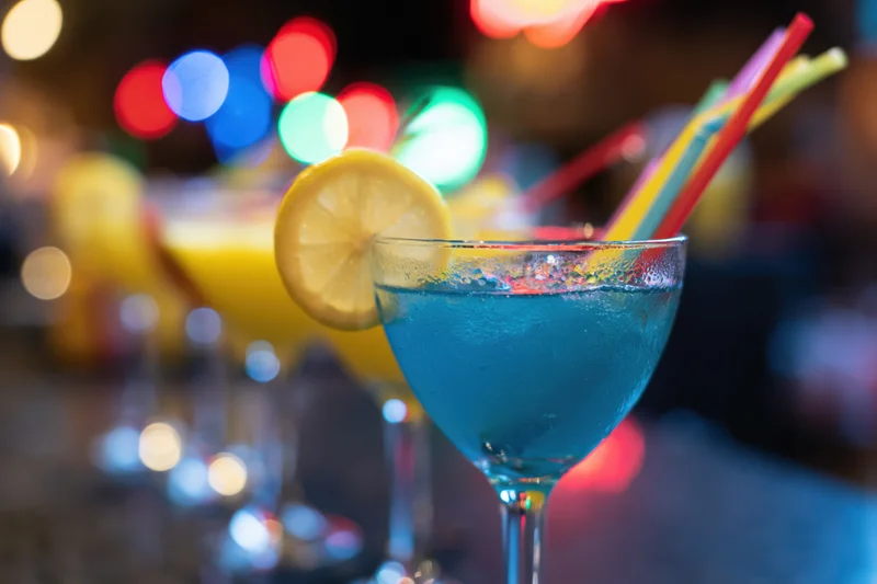 Blue cocktail in a glass with lemon slice and rainbow straw on a bar counter at night