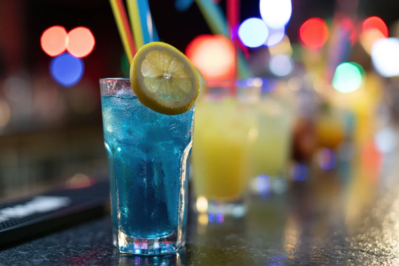 Blue cocktail with lemon garnish and straw on dark bar counter at night