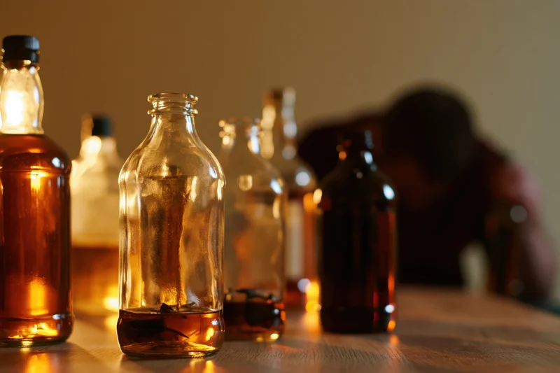 Empty and half-full glass liquor bottles arranged on a wooden table with a blurred man in the background under warm dim lighting