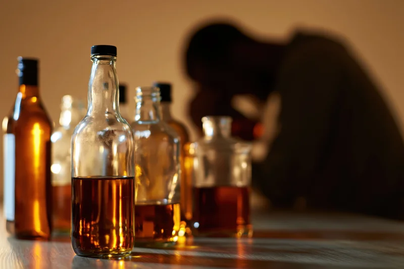 Several glass liquor bottles on a wooden table with a blurred silhouette of a person in distress in the background