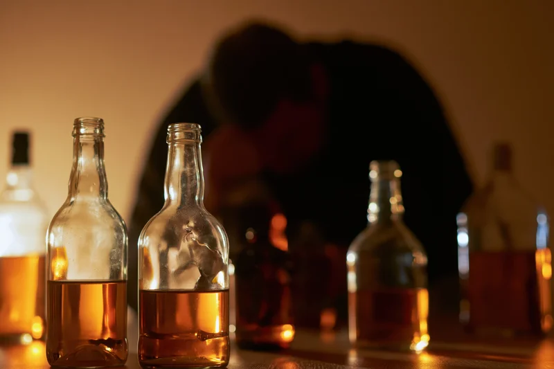 Empty and half-full whiskey bottles on a dimly lit table with a blurred silhouette of a man in the background