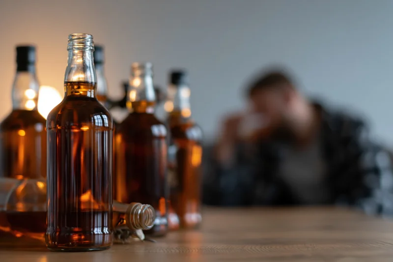 Several glass alcohol bottles arranged on a wooden table with a blurred man in the background