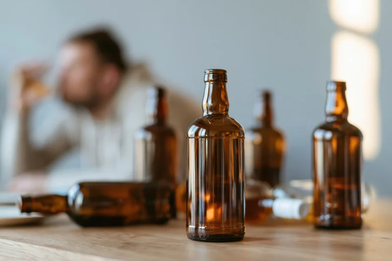 Empty brown glass alcohol bottles arranged on wooden table with blurred man drinking in background