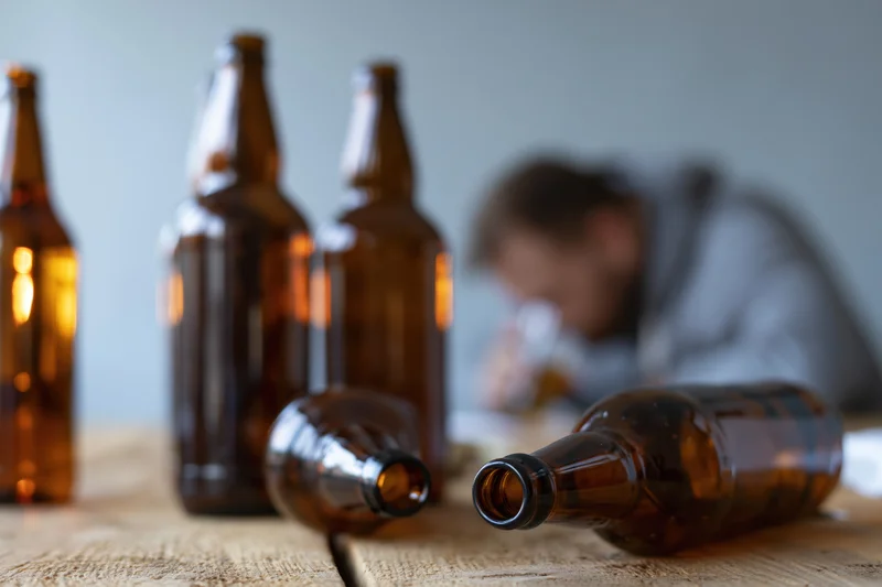 Empty beer bottles on a wooden table with a blurred man drinking in the background