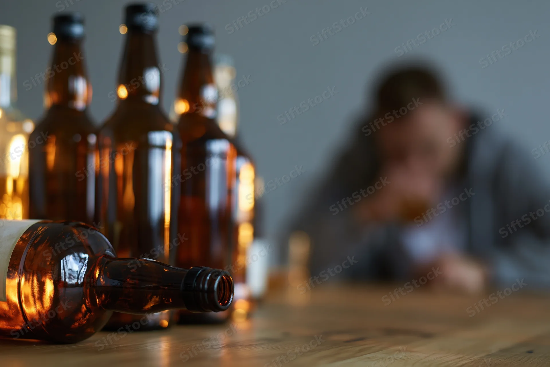 Several glass alcohol bottles on a wooden table with a blurred man drinking in the background