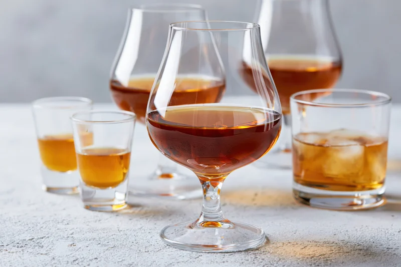 Whiskey and cognac glasses arranged on a light grey surface with ice and shot glasses nearby