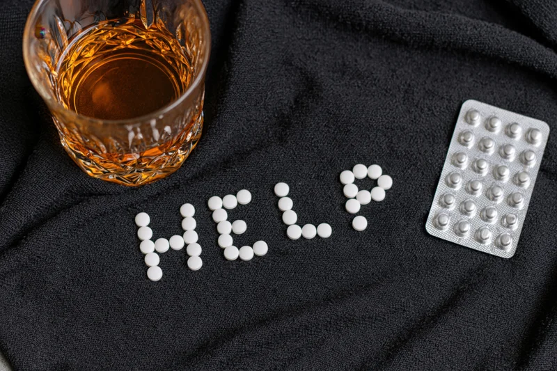 White pills arranged to spell help on dark fabric beside a glass of whiskey and a blister pack