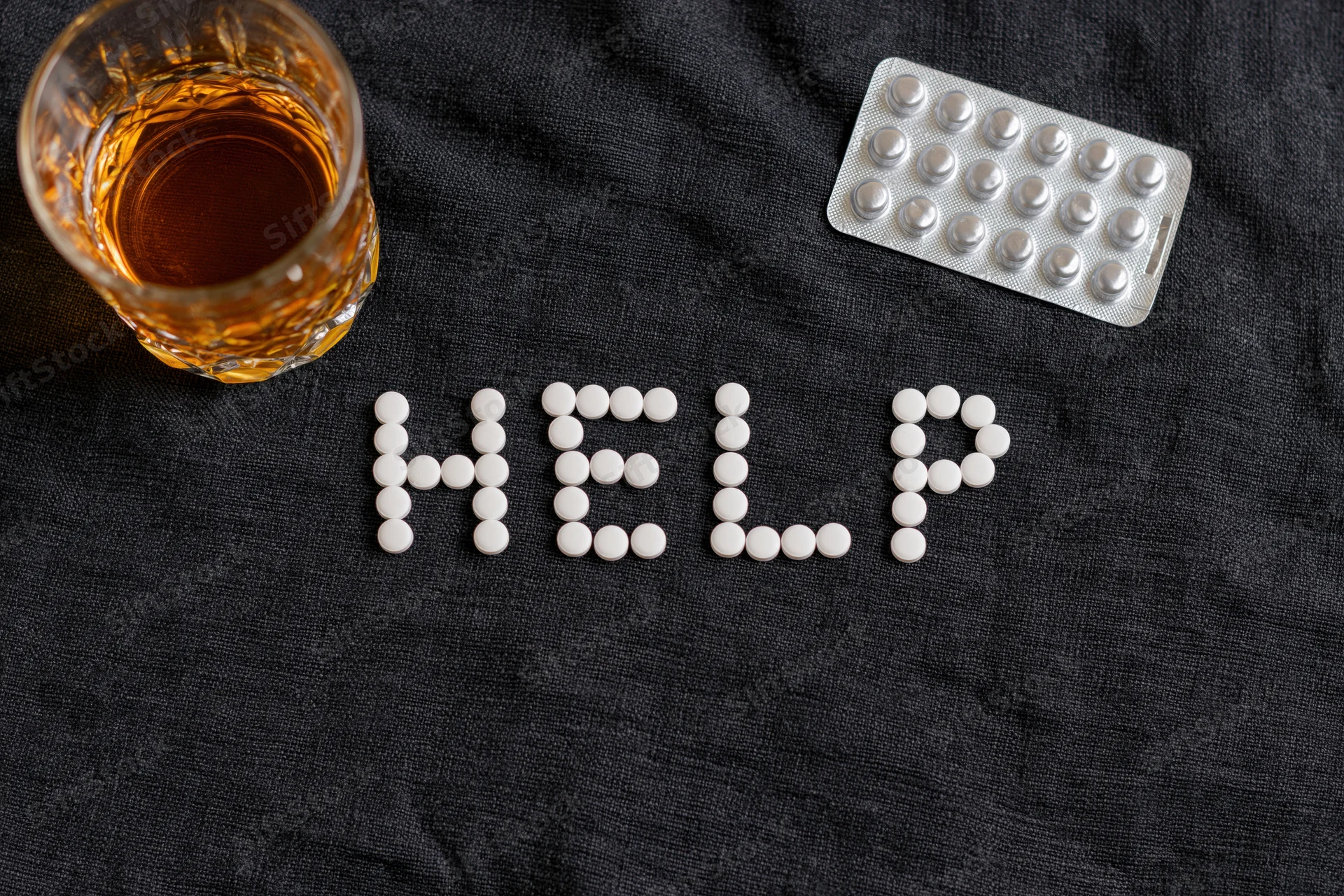 White pills arranged to spell help on dark fabric beside a glass of whiskey and a blister pack