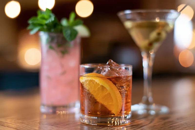 Three cocktails arranged on a wooden bar top with garnishes and ice under warm dim lighting