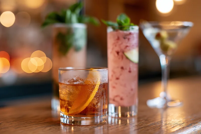 Three cocktails with garnishes arranged on a wooden bar top under warm ambient lighting