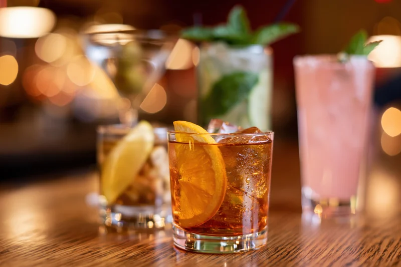 Several cocktails with garnishes arranged on a wooden bar top in a dimly lit lounge