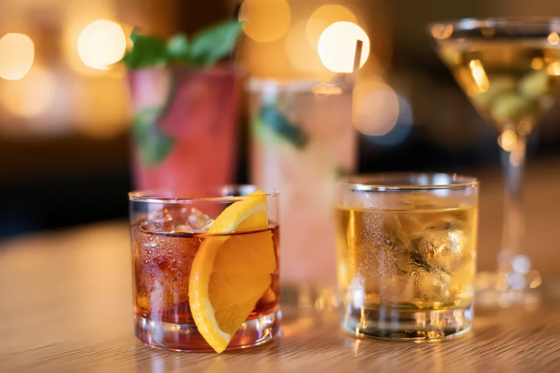 Three cocktails with garnishes arranged on a wooden bar top under warm ambient lighting