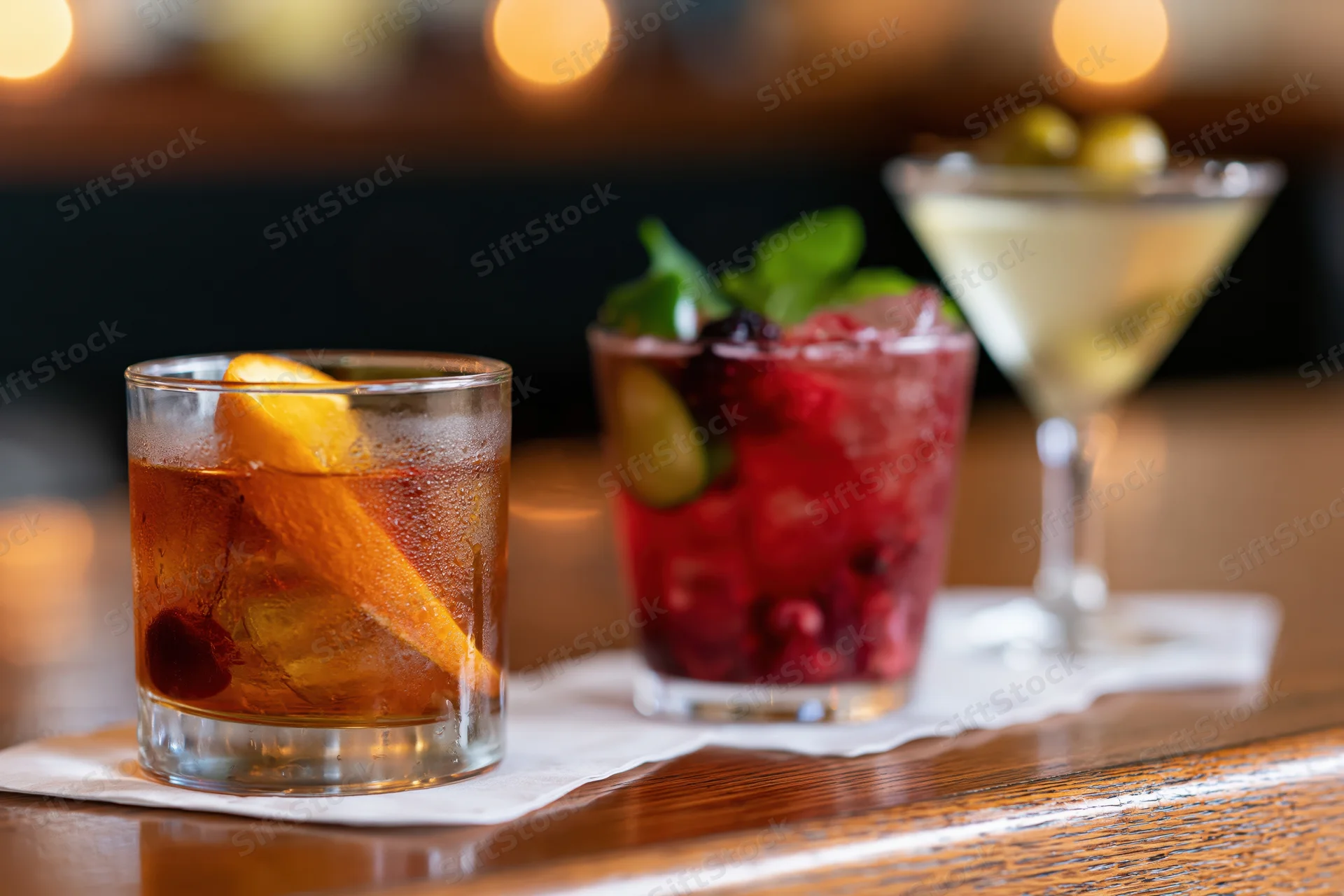 Three cocktails with garnishes arranged on a wooden bar top under warm lighting